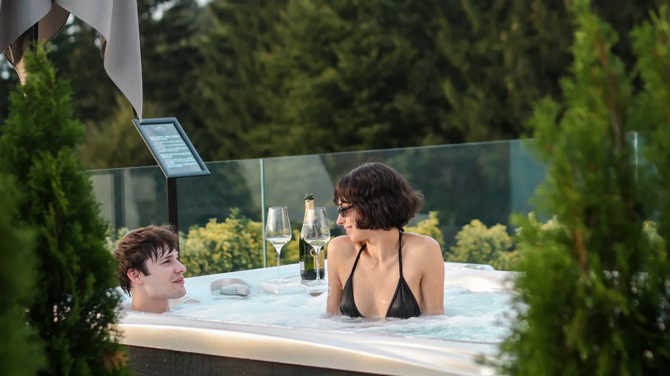 Couple in an outdoor Eden Hill hot tub, with a bottle and glasses on the edge and forest behind them.