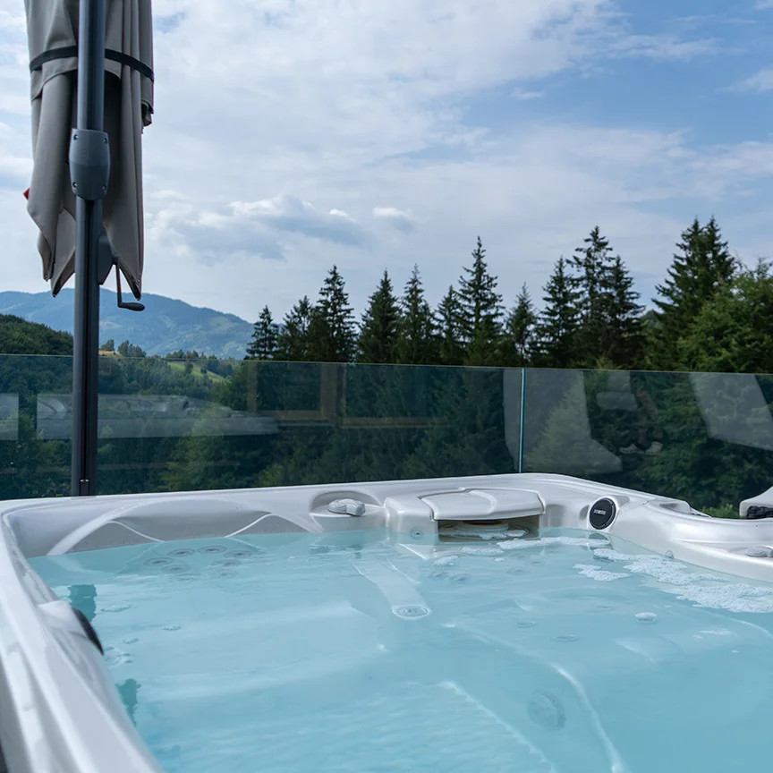 Hot tub on an Eden Hill terrace with a glass railing, overlooking fir trees and mountains.