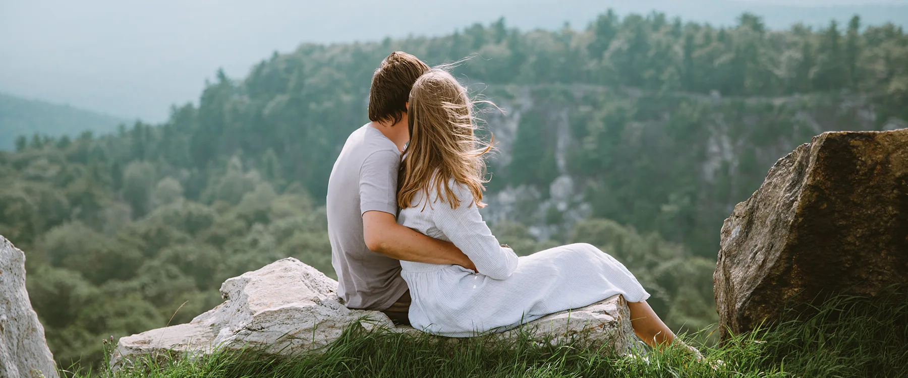 Couple embracing on a rocky outcrop overlooking forested hills.