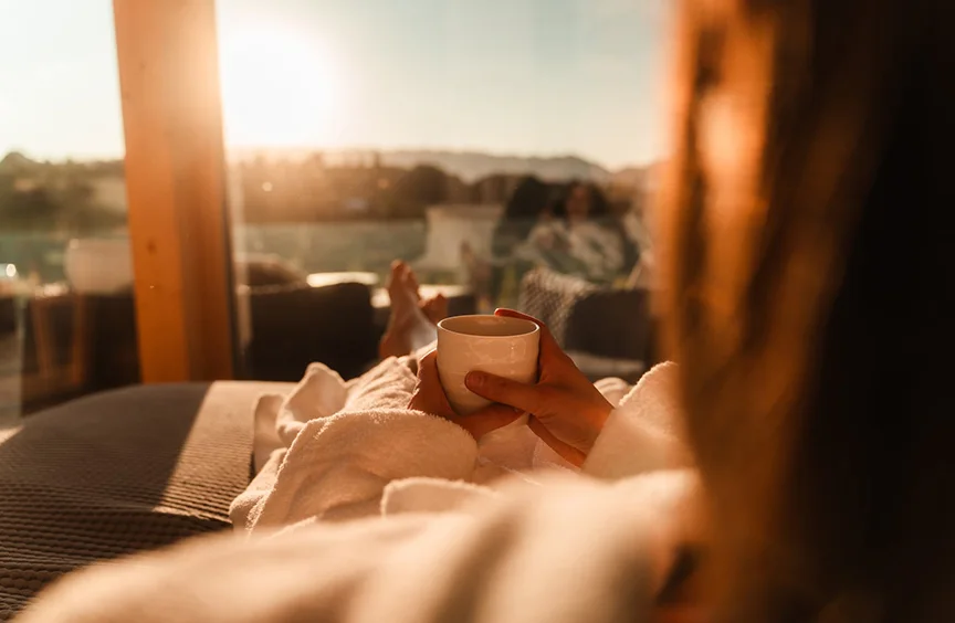 Person wrapped in a blanket, holding a white mug beside a window in the sunset light.