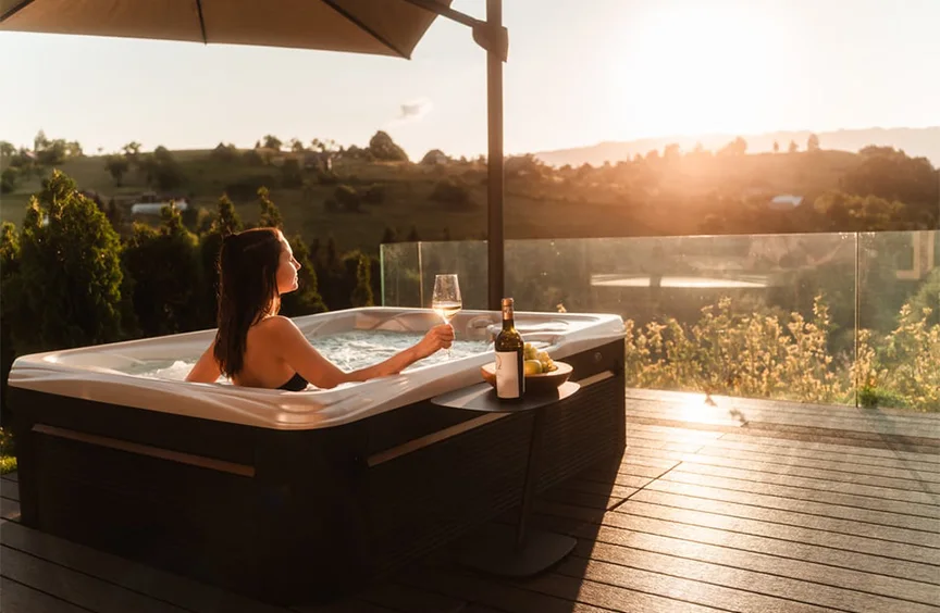Woman in an outdoor Eden Hill hot tub at sunset, with a glass and bottle on the table beside her.