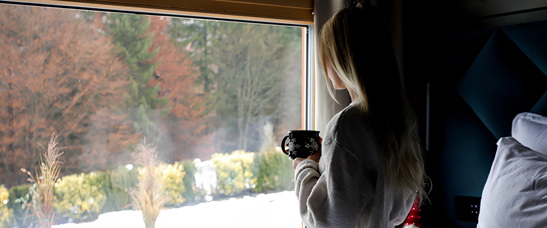Woman holding a mug beside an Eden Hill bedroom window, looking out at the snow.
