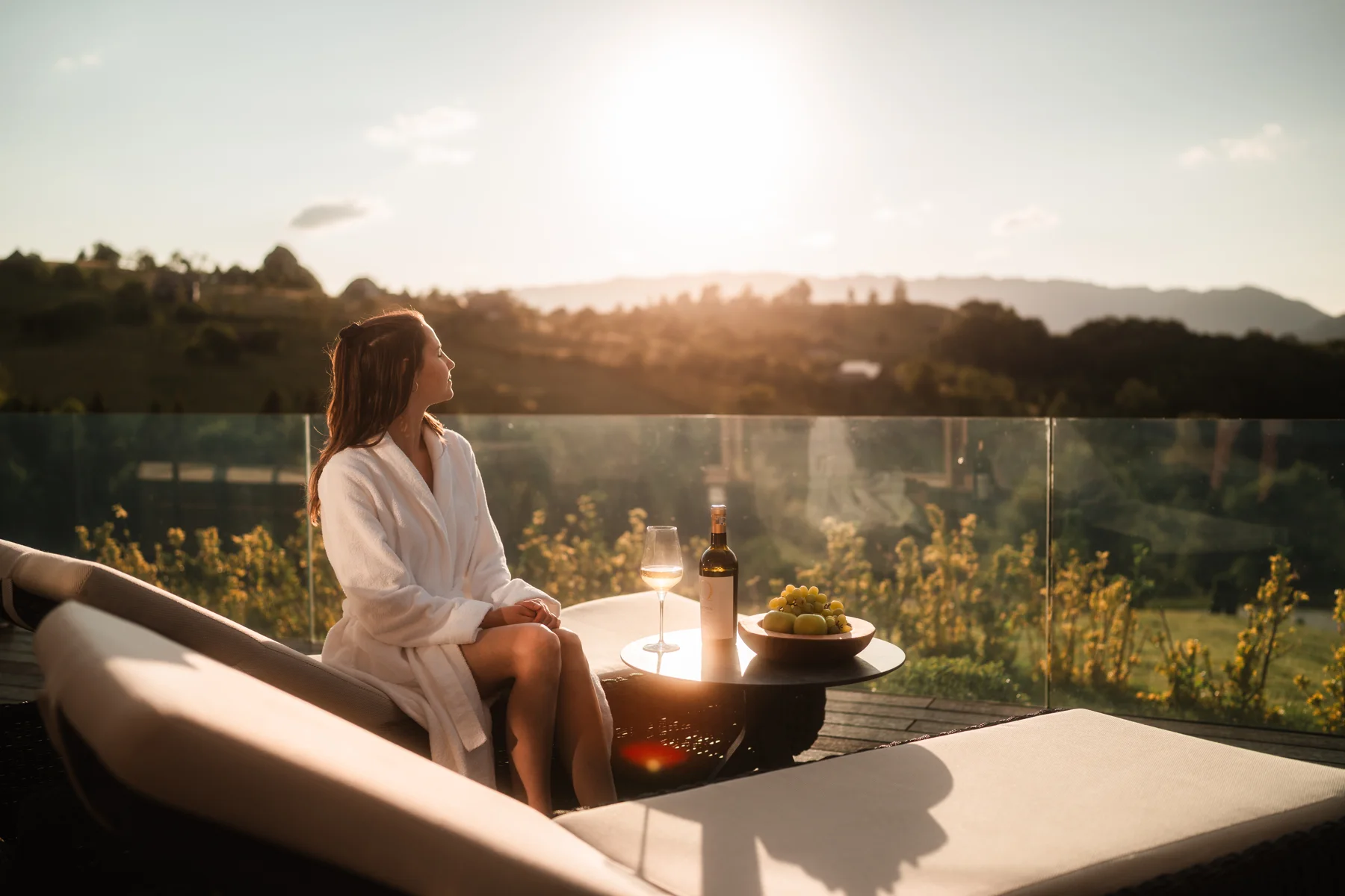 Woman in a white robe on an Eden Hill terrace beside a table with wine and grapes at sunset.