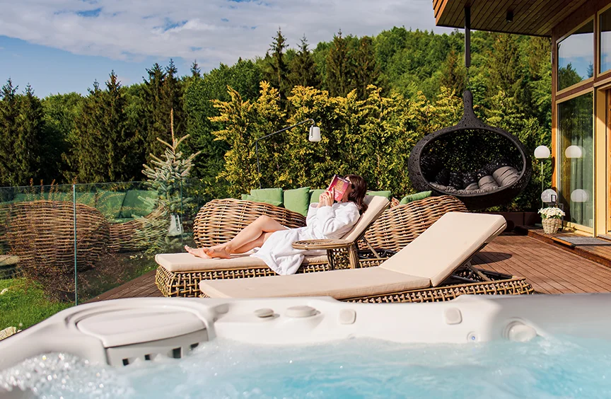 Woman on a wicker lounger beside an Eden Hill hot tub, with a hanging chair and forest behind her.
