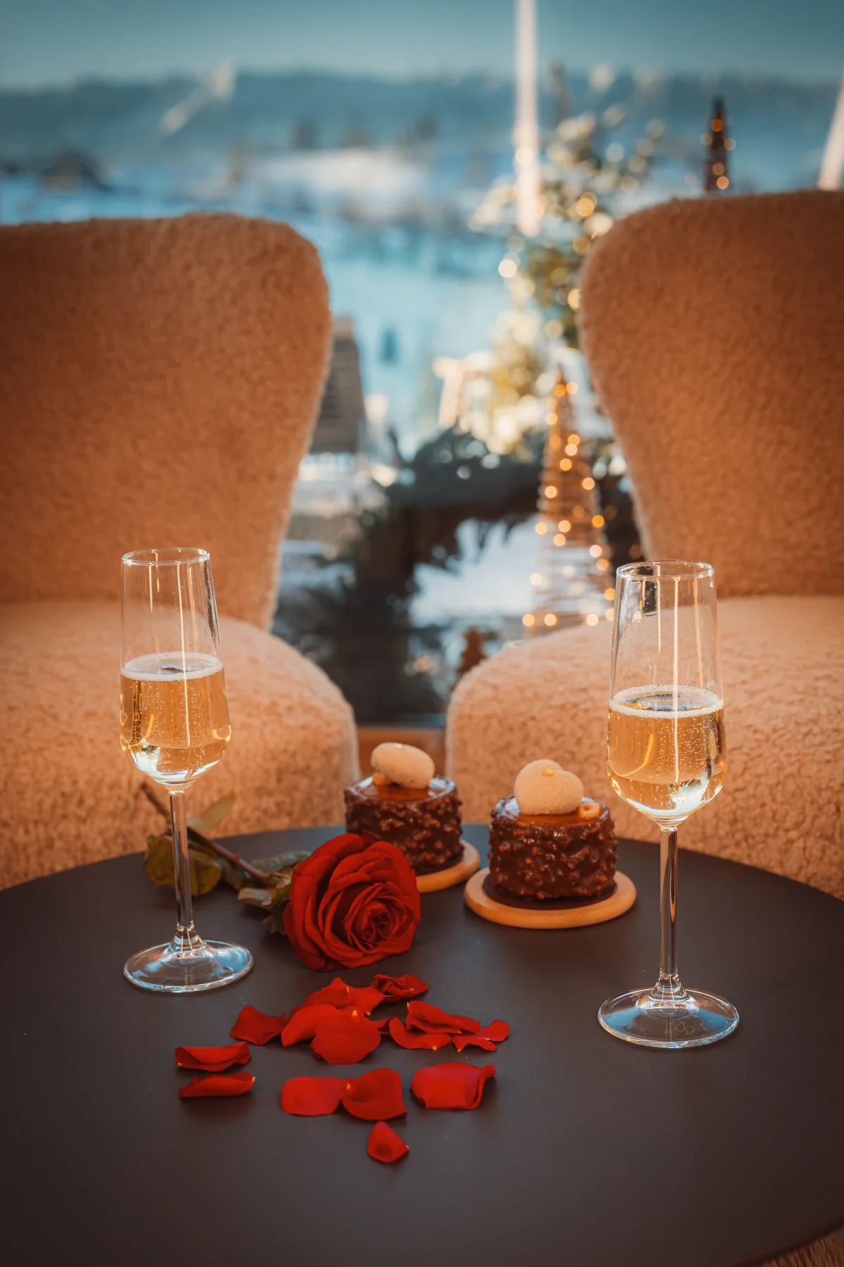 Two glasses of sparkling wine, desserts and a rose on a small table beside a window.