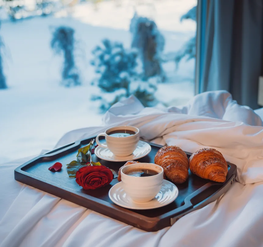 Tray with two coffees, croissants and a rose on a white bed beside a window overlooking a winter landscape.