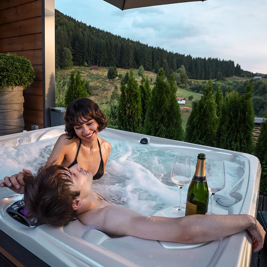 Couple in an Eden Hill hot tub, with a bottle on the edge and forested hills behind them.