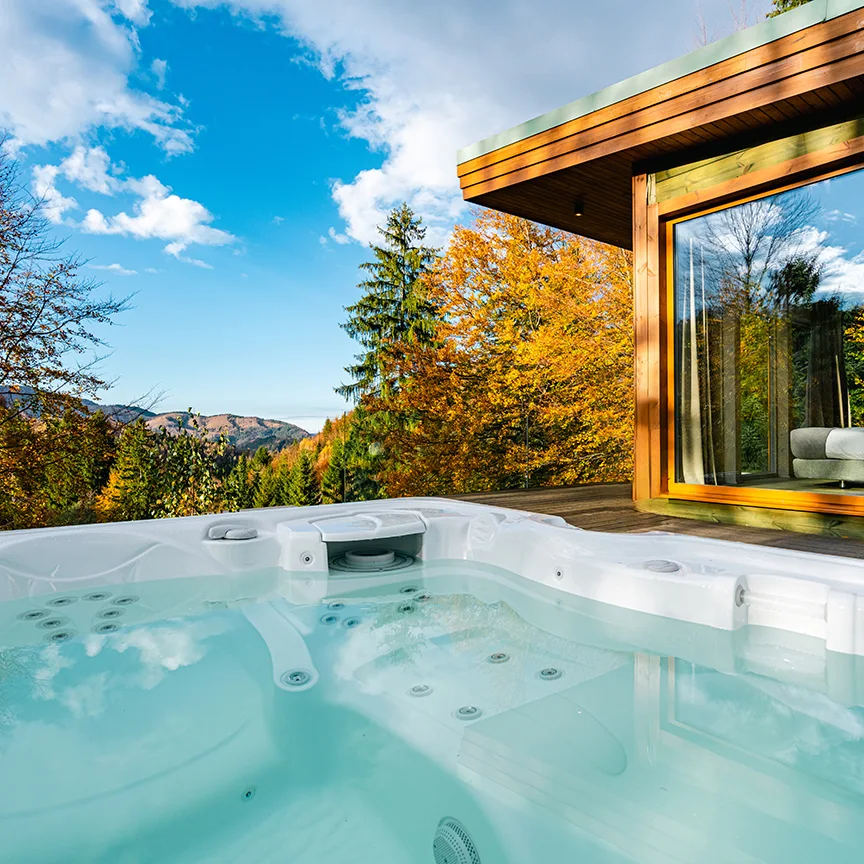 Outdoor hot tub beside a wooden Eden Hill villa, with an orange autumn tree beneath a blue sky.