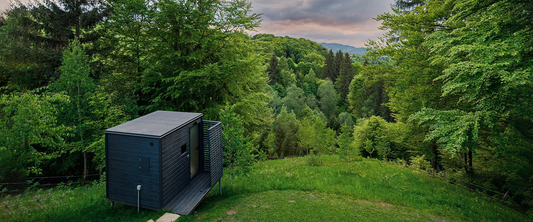 Wooden Eden Hill sauna at the edge of a green clearing, with forest and a sunset sky behind it.