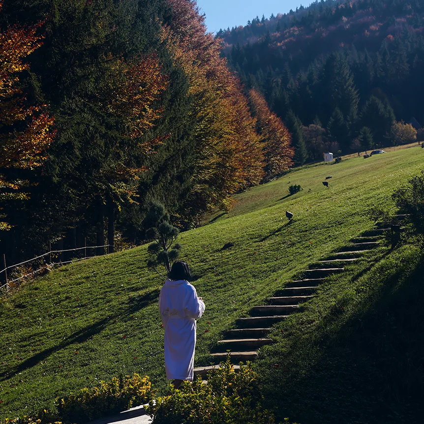 Person in a white robe walking uphill beside the Eden Hill garden steps among autumn trees.