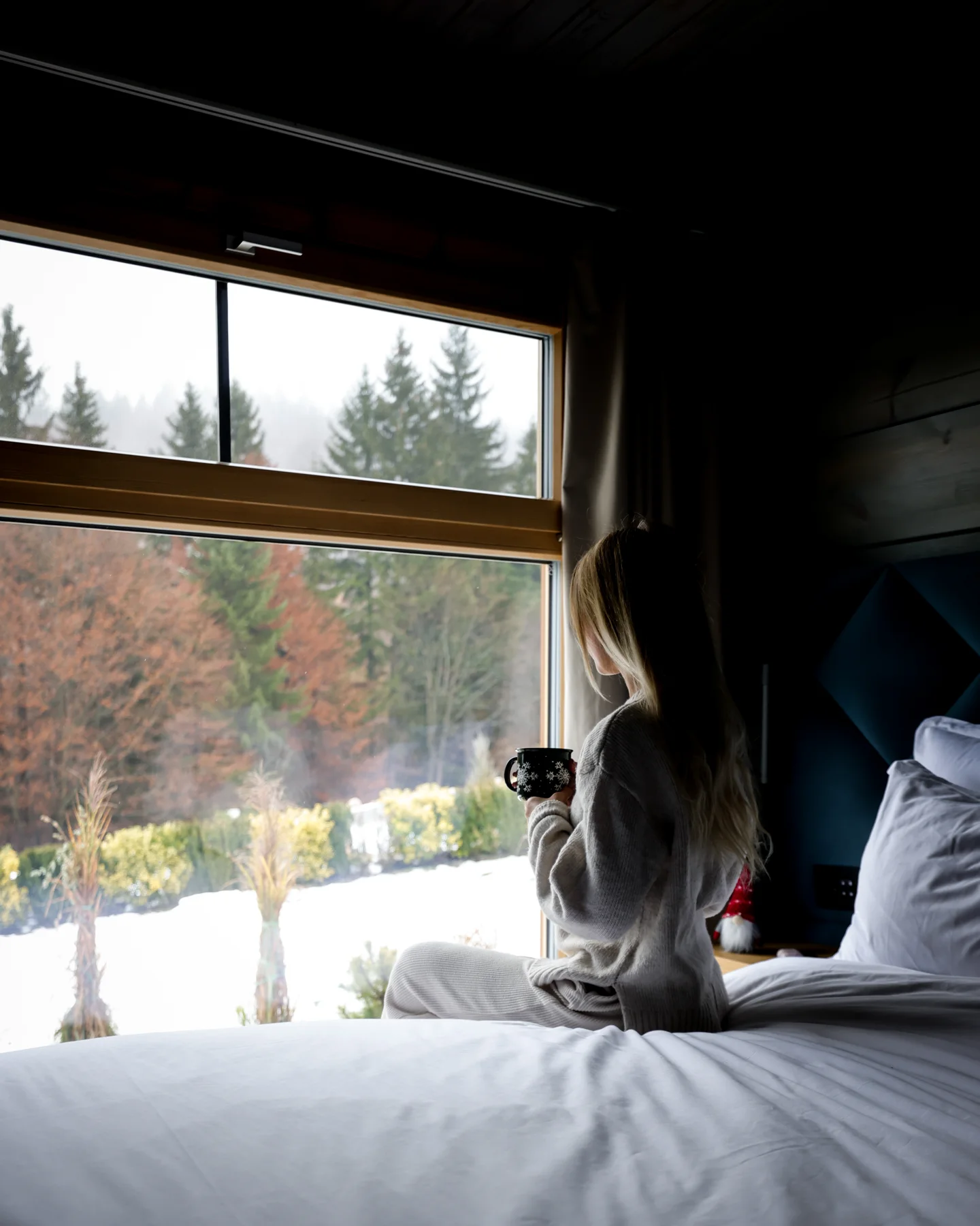 Woman sitting on a bed holding a mug beside a large window overlooking the snowy Eden Hill garden.
