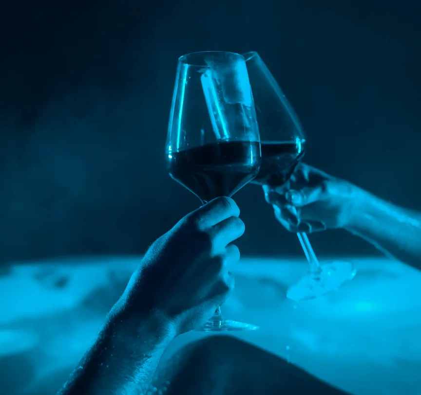 Two wine glasses clinking above a bubbling, blue-lit hot tub.