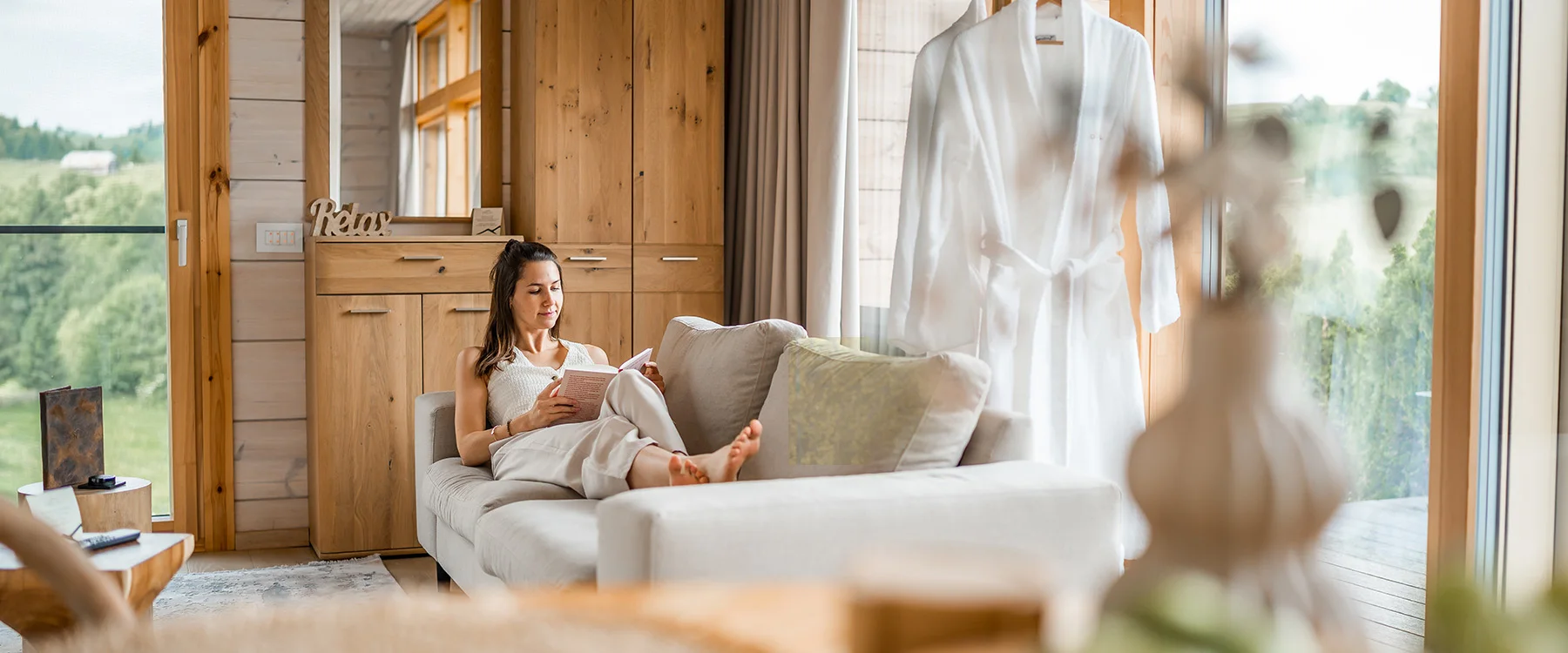 Woman reading on a cream sofa in an Eden Hill living room beside large windows and wooden walls.
