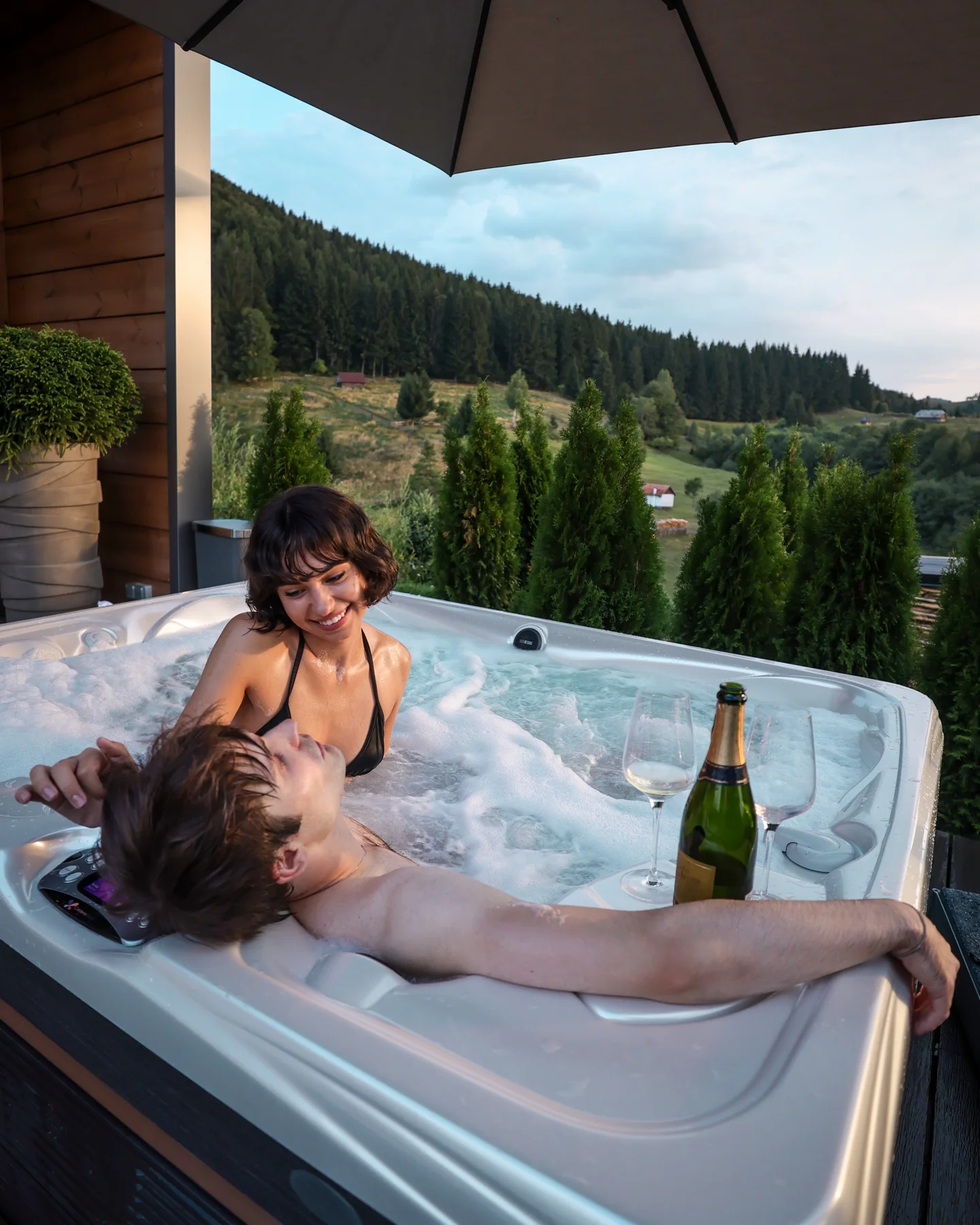Couple in a hot tub on a covered Eden Hill terrace, with a bottle on the edge and forest behind them.